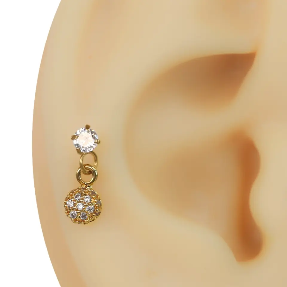 Kulak tragus ve helix bölgesinde sergilenen zirkon taşlı sallantılı şans topu piercing tasarımı.