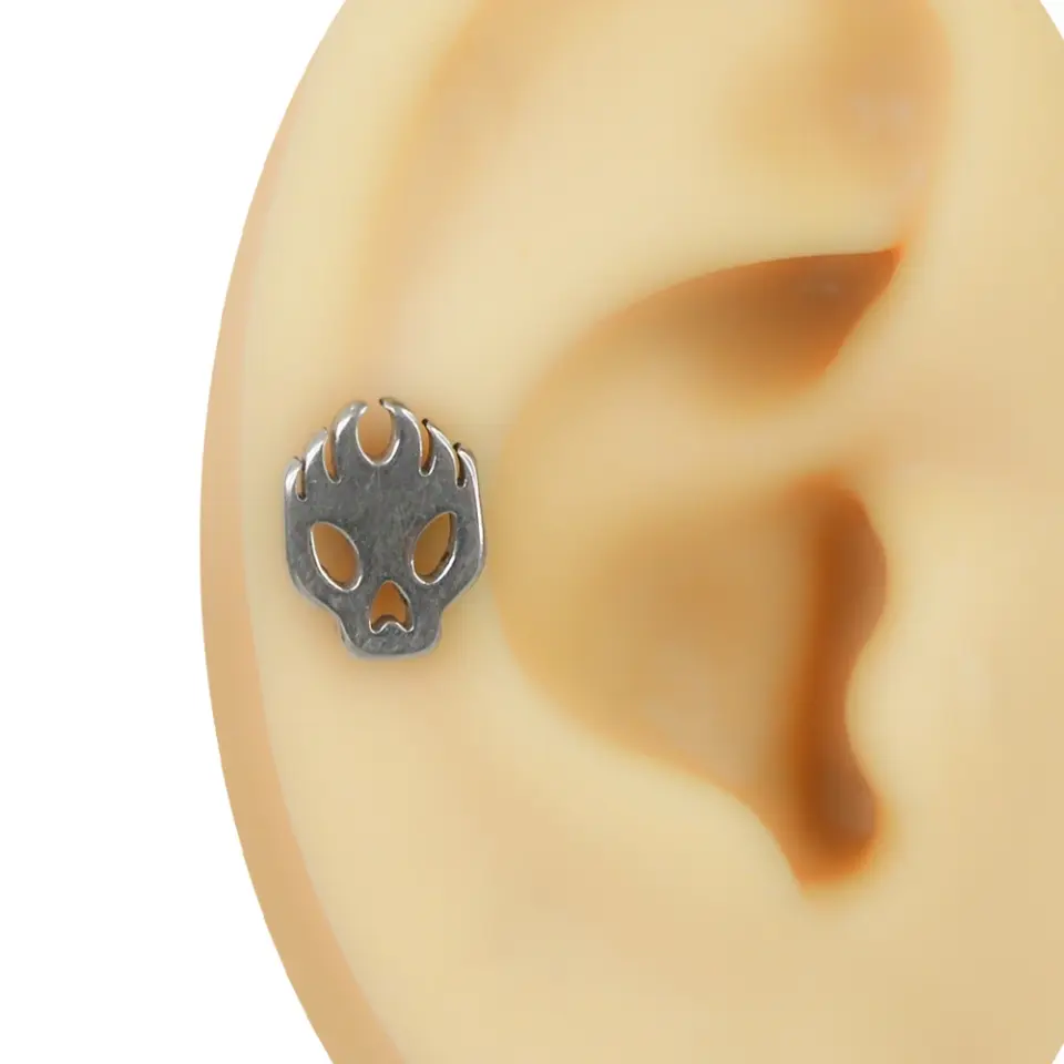 Kulakta helix kıkırdak bölgesinde takılı gümüş renkli alev detaylı kurukafa figürlü flatback piercing
